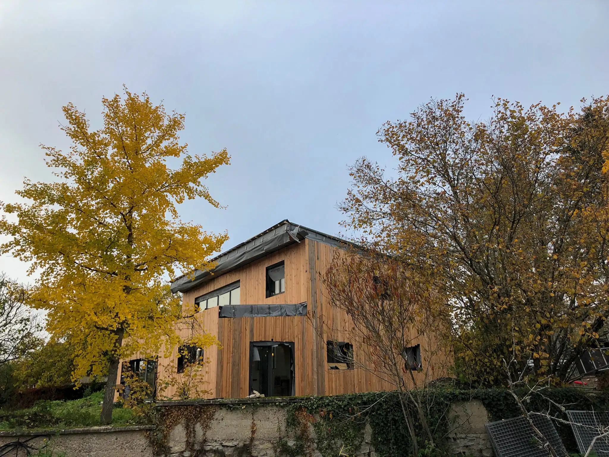 A l'arrière plan la maison en bois-paille, avec un bardage bois vertical posé sur les murs. Au premier plan, de la végétation