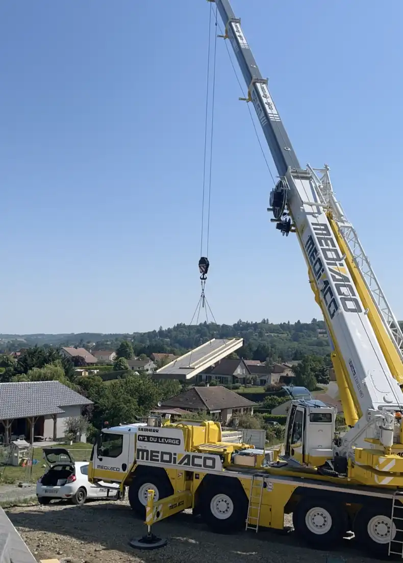 Grue au premier plan en train de décharger un caisson de toiture isolé en paille. Le caisson est directement emmené du camion pour être posé sur la maison.