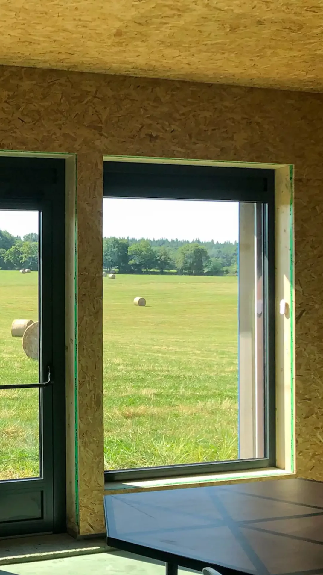 Photo depuis l'intérieur d'une salle en bois paille, on aperçoit à travers les ouvertures un champ avec des bottes de paille. L'intérieur de la salle a une finition brute en OSB.